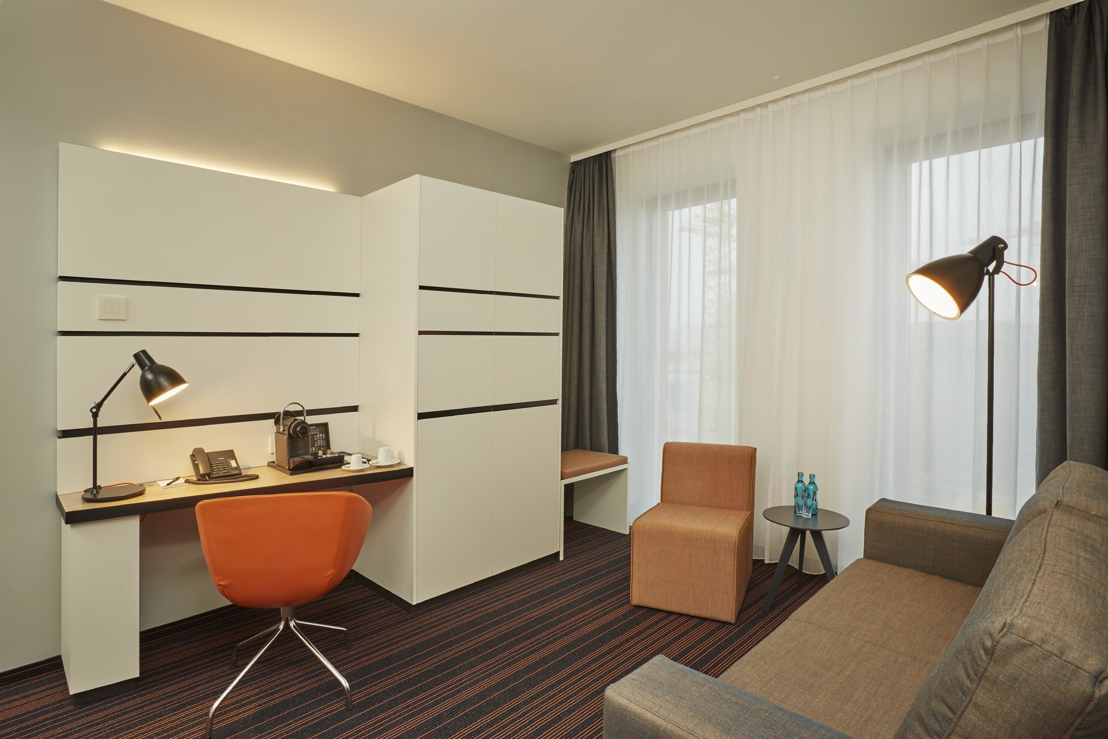 A hotel room with a desk, a chair, a sofa, a lamp, and a window with curtains.