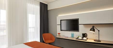 A modern hotel room with a large bed, a desk with a flat-screen TV, a chair, and a desk lamp.