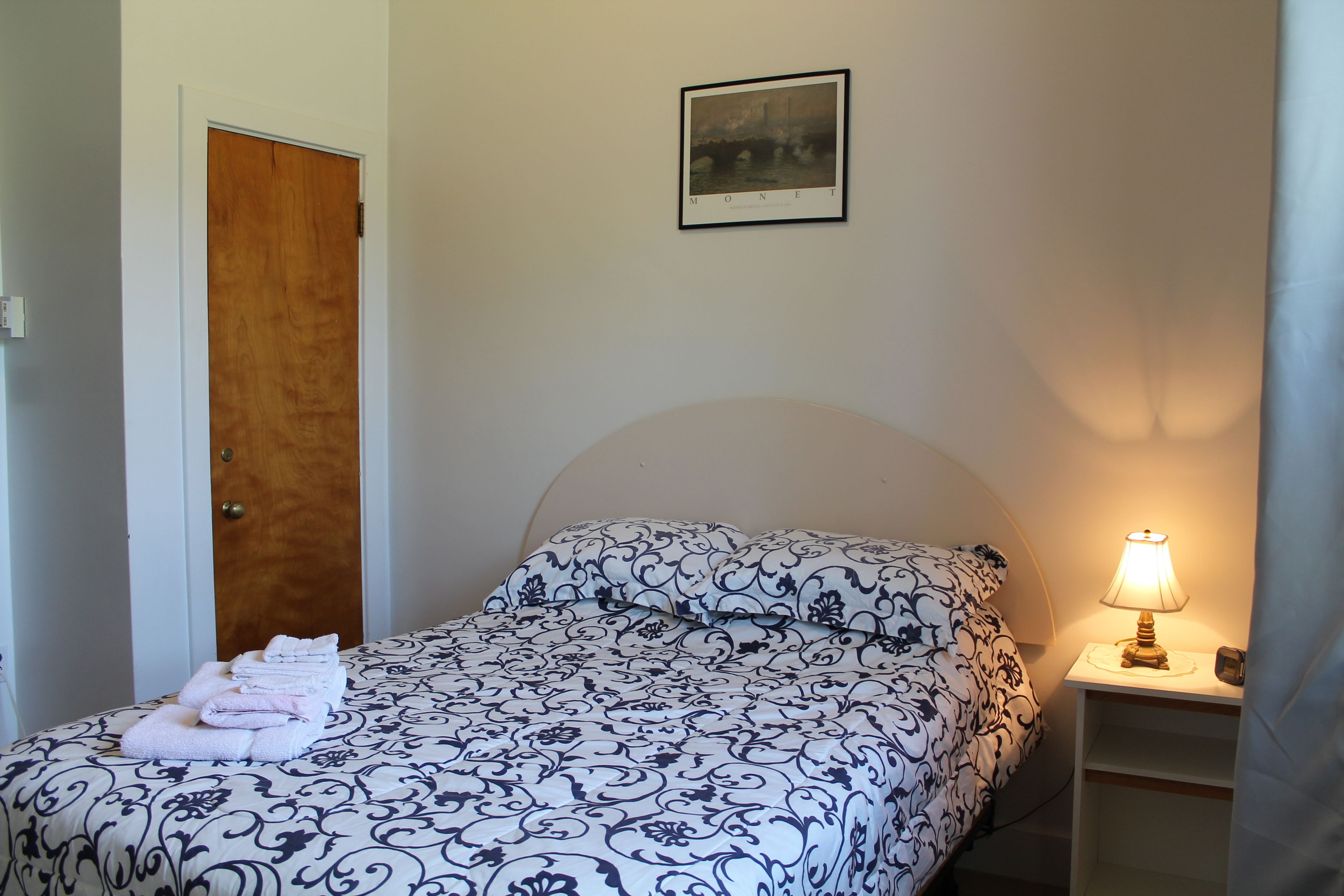 A bedroom with a bed, a nightstand with a lamp, a door, and a framed picture on the wall.
