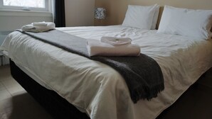 Una cama bien hecha con sábanas blancas y una manta gris, una ventana con vistas a árboles desnudos y una lámpara de noche.