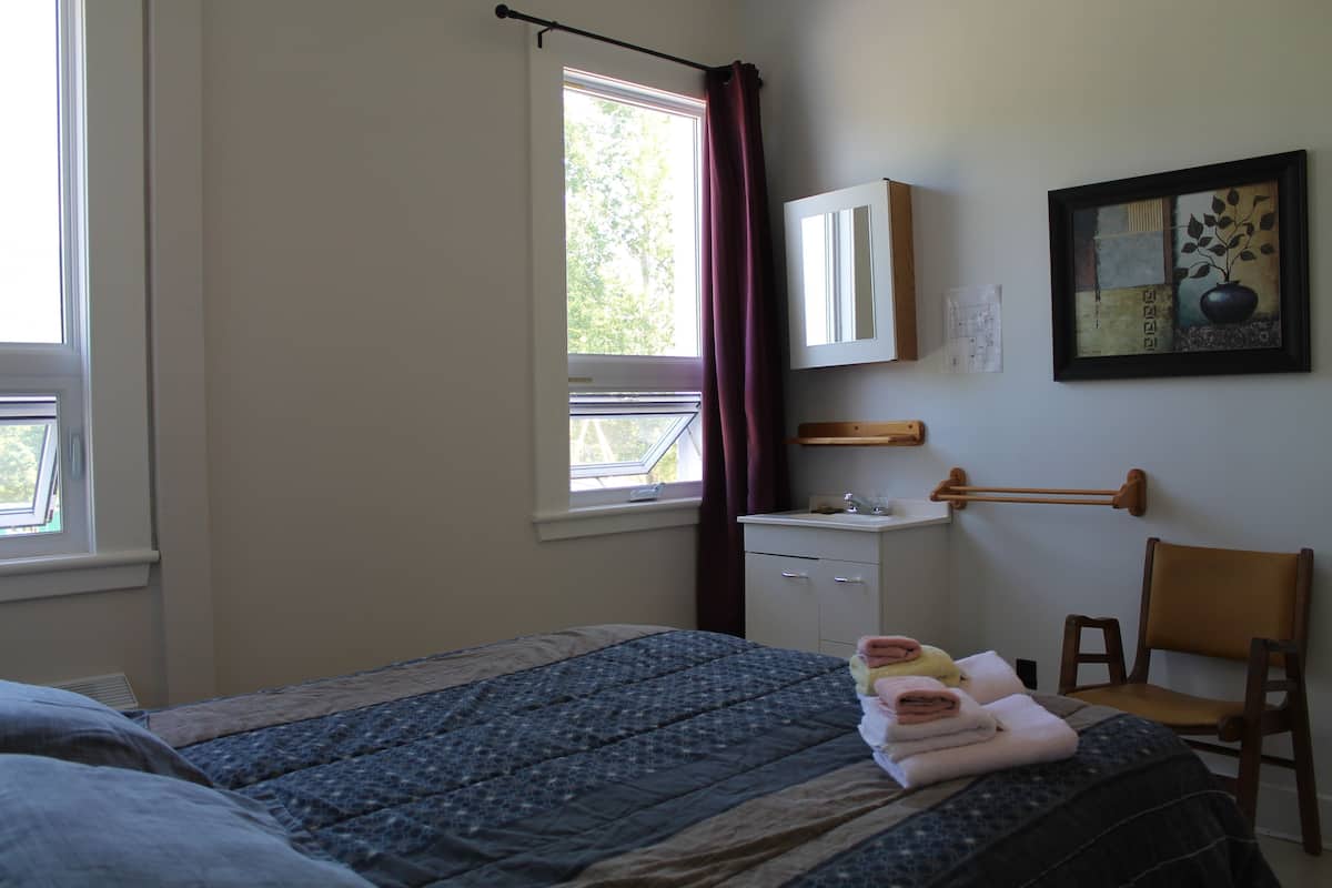 Un dormitorio con cama, una silla, una ventana con cortinas, un escritorio pequeño y un espejo montado en la pared.