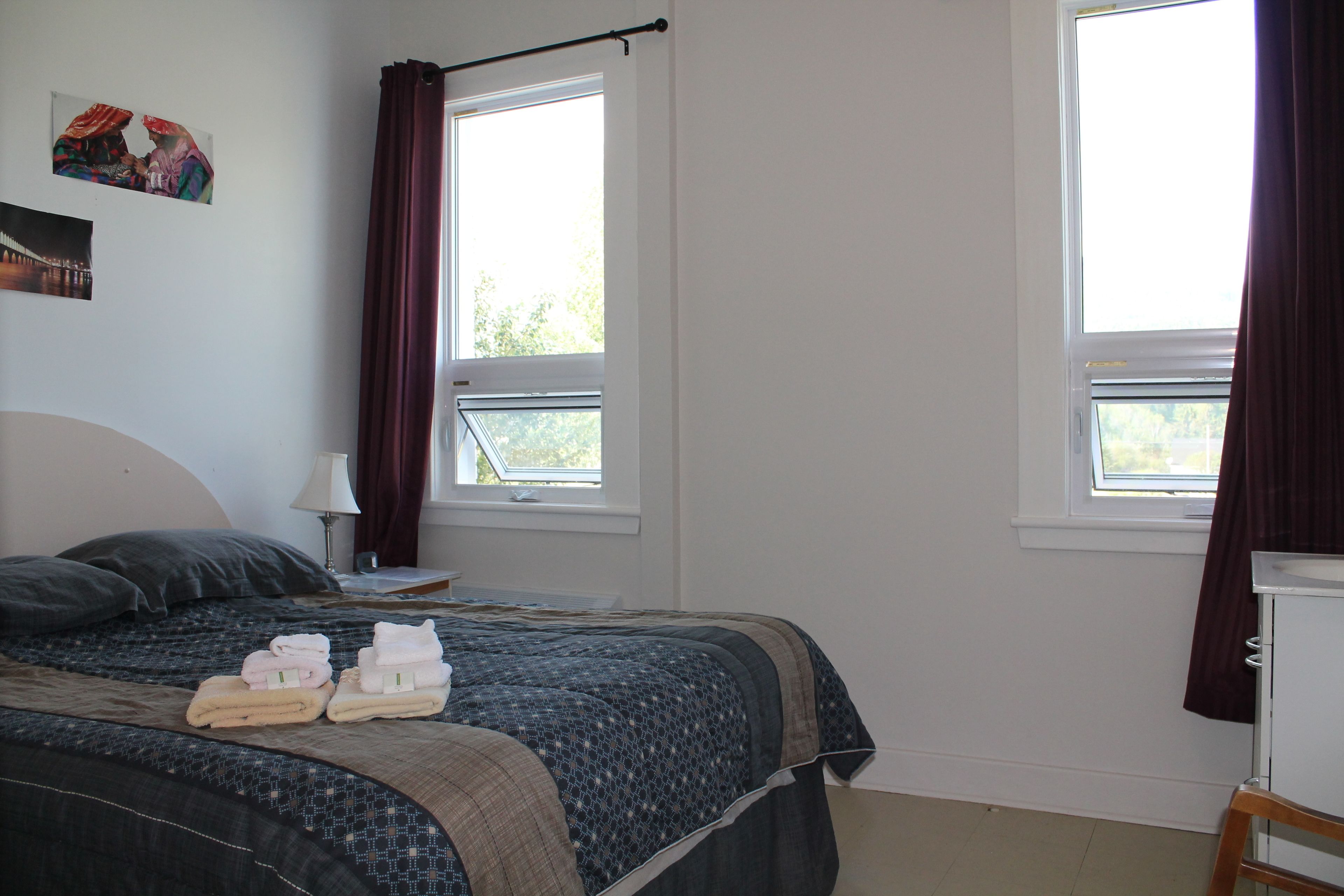 Une chambre à coucher avec un lit, deux fenêtres, une table de chevet, une lampe et un tableau accroché au mur.