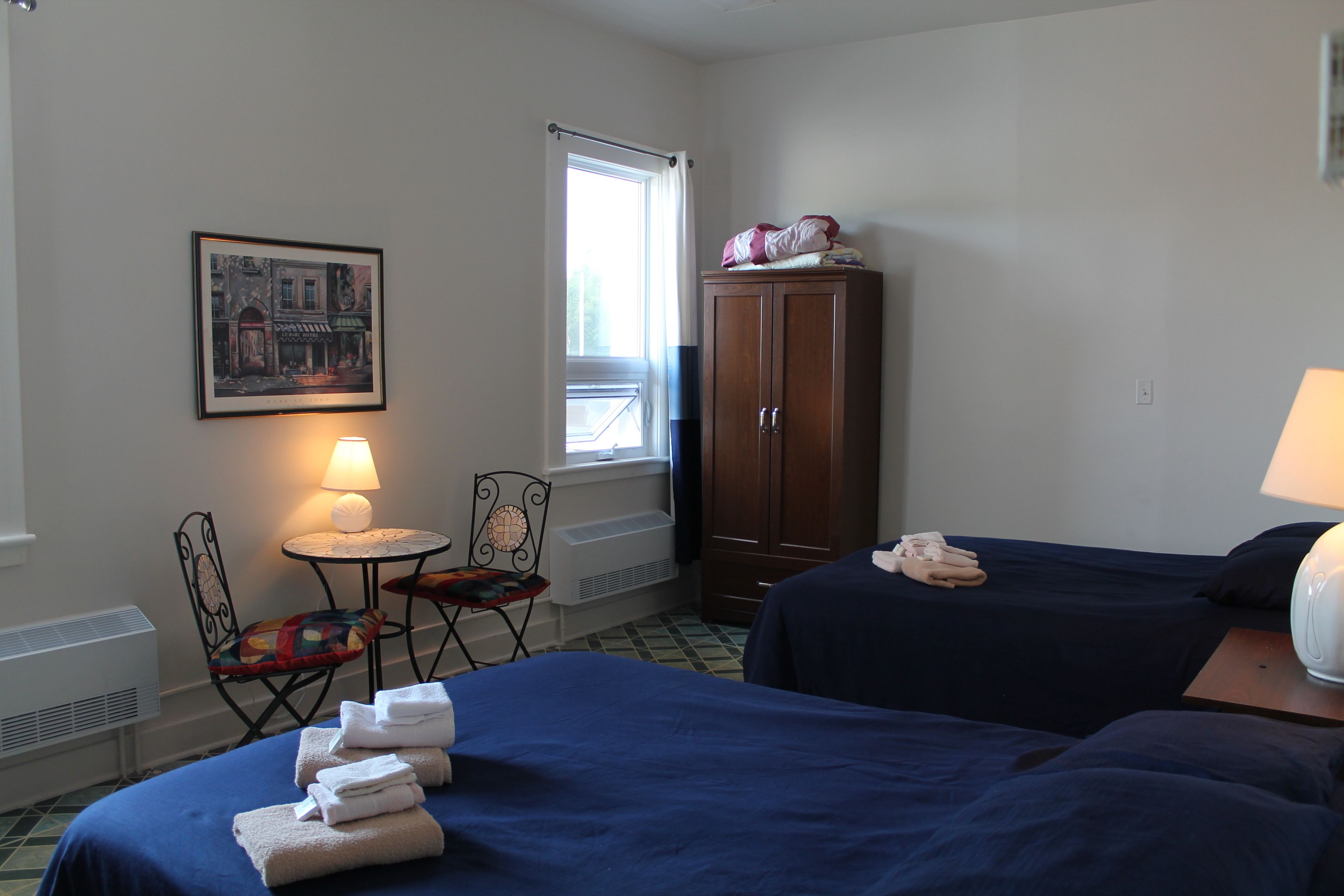 Habitación de hotel con dos camas, una silla, una mesita con una lámpara, un armario y un cuadro en la pared.