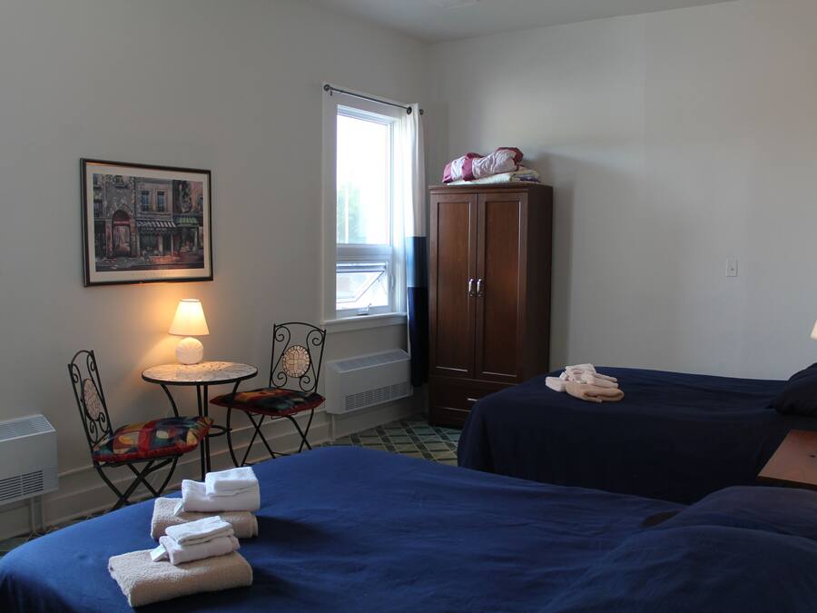 Habitación de hotel con dos camas, una silla, una mesita con una lámpara, un armario y un cuadro en la pared.