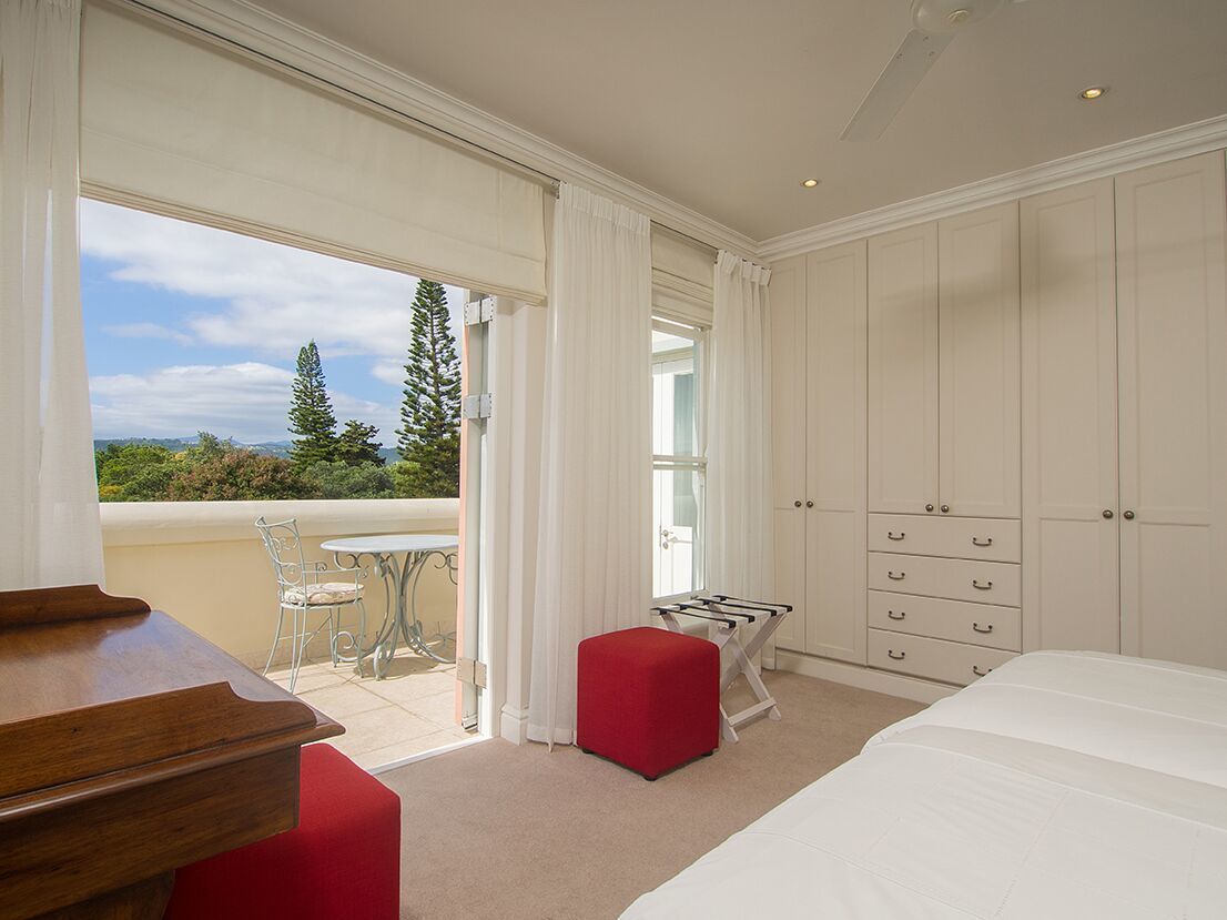 A bedroom with a large bed, a wooden dresser, a red ottoman, and a balcony with a table and chairs.