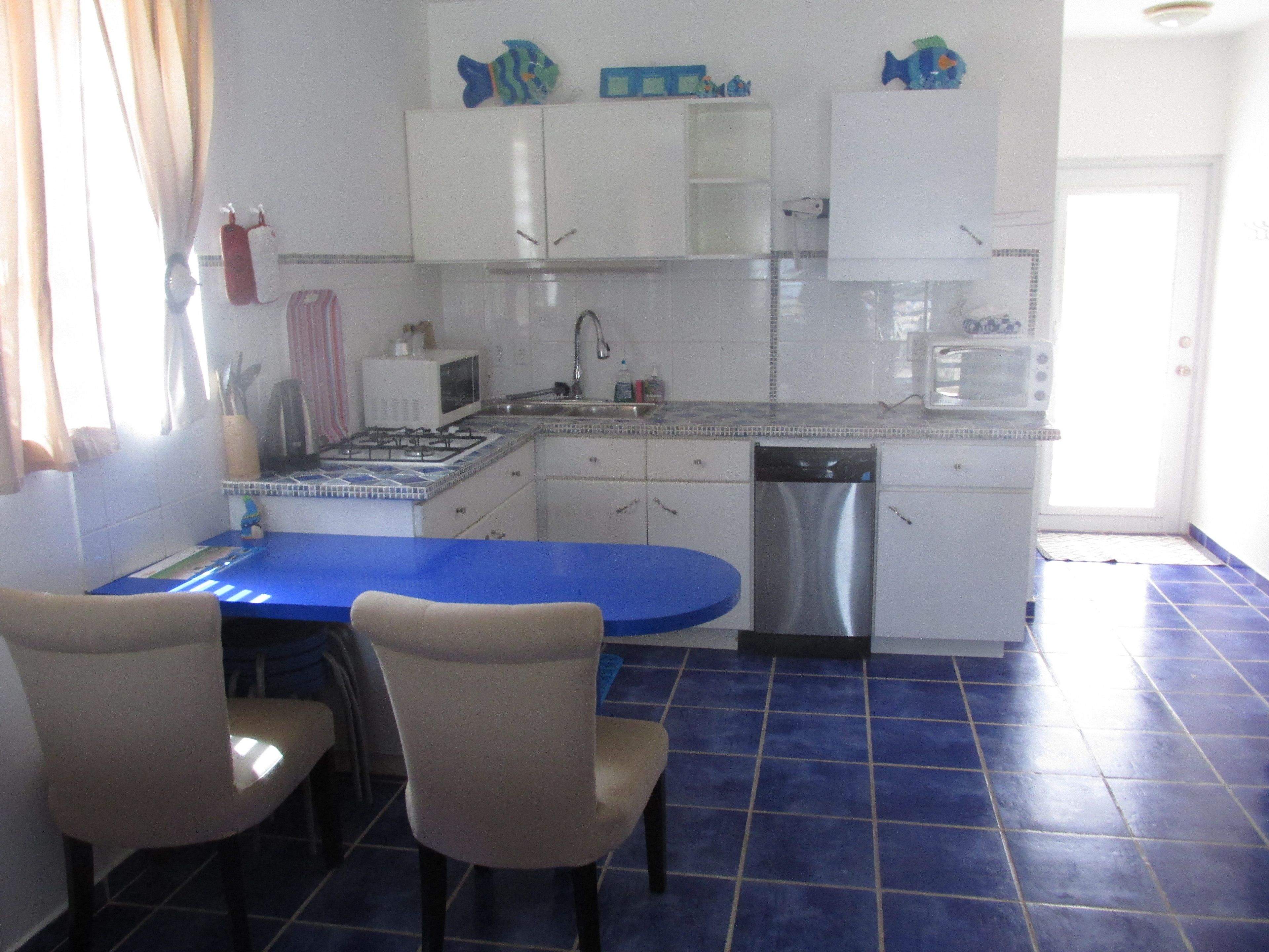 Una cocina con encimera azul, armarios blancos y lavavajillas. Hay dos sillas en la mesa.