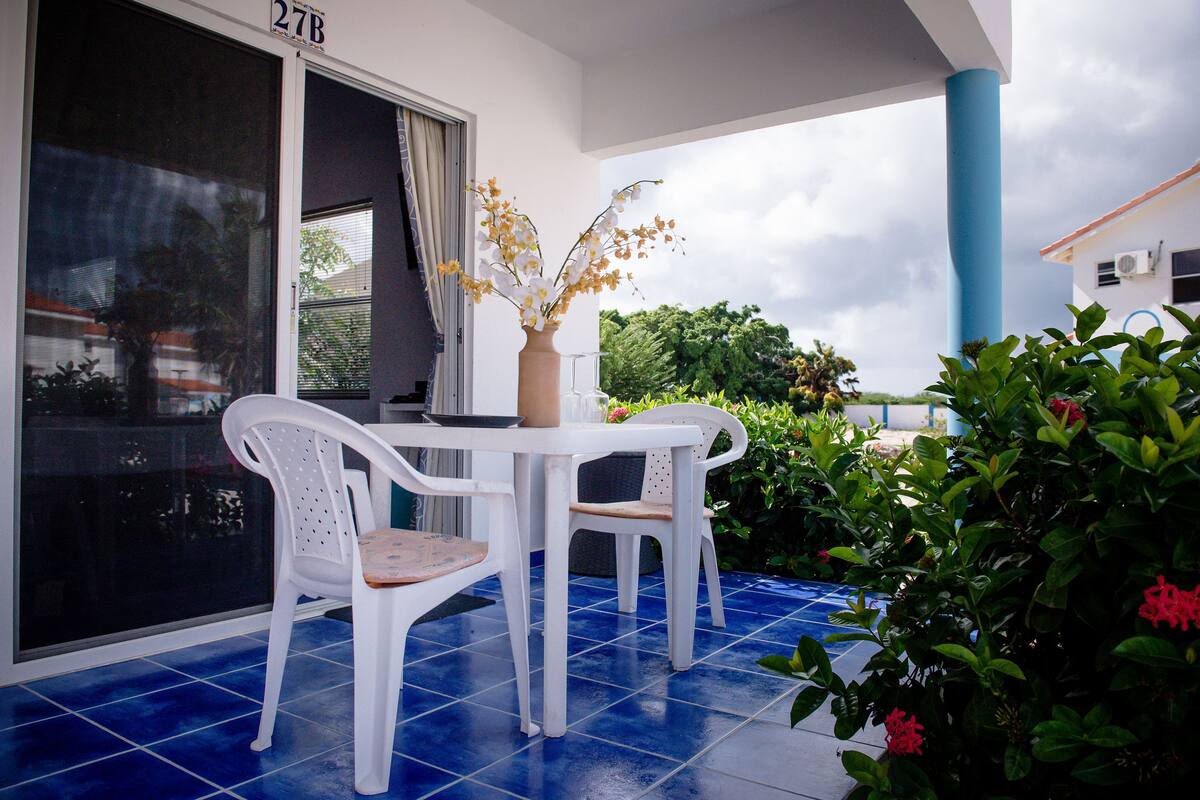 Ein Balkon mit einem weißen Tisch und zwei Stühlen, einer Blumenvase und Blick auf Grünflächen und ein Gebäude.