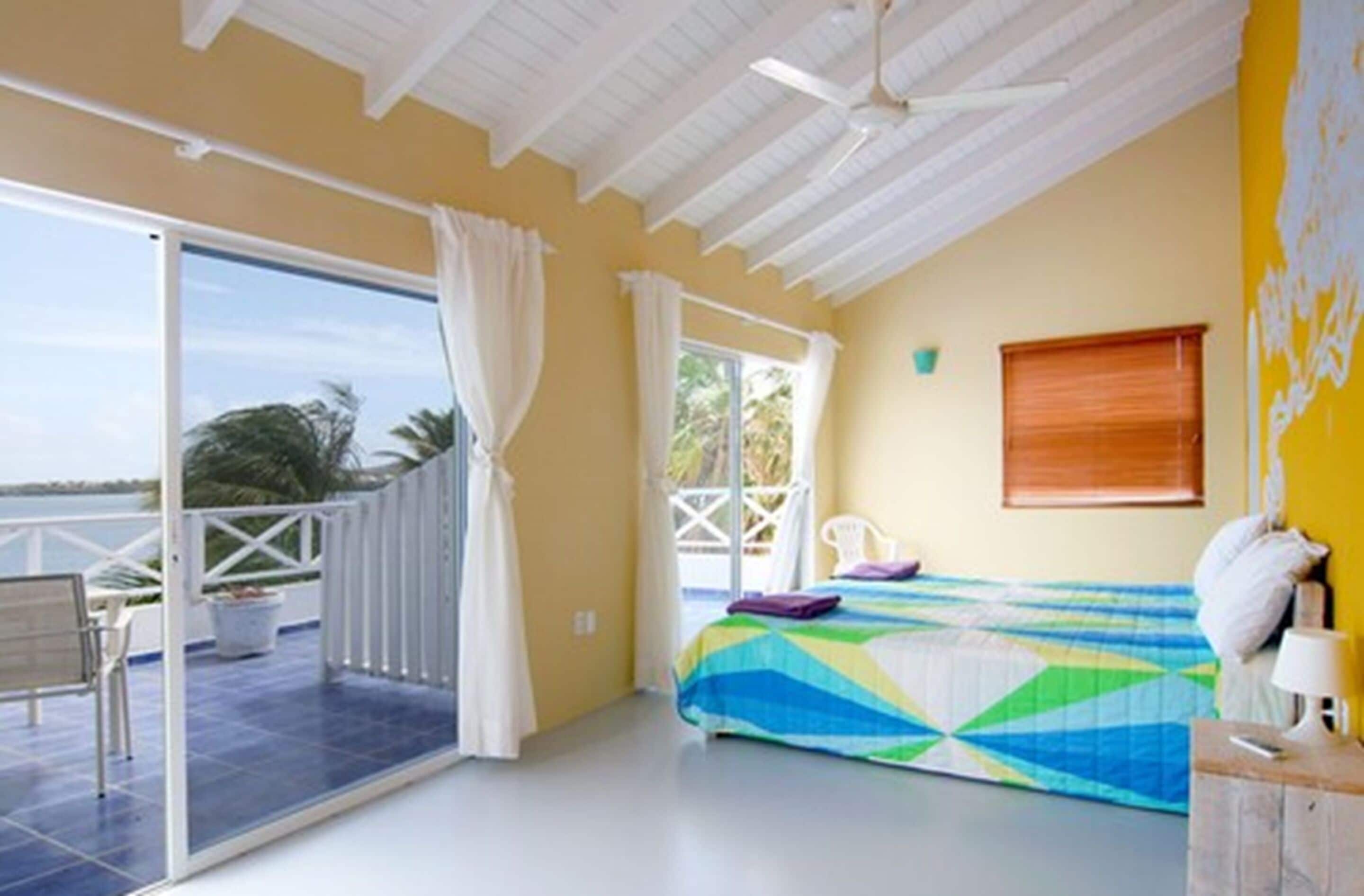 A room with a bed, a window with curtains, a ceiling fan, and a view of the outdoors.