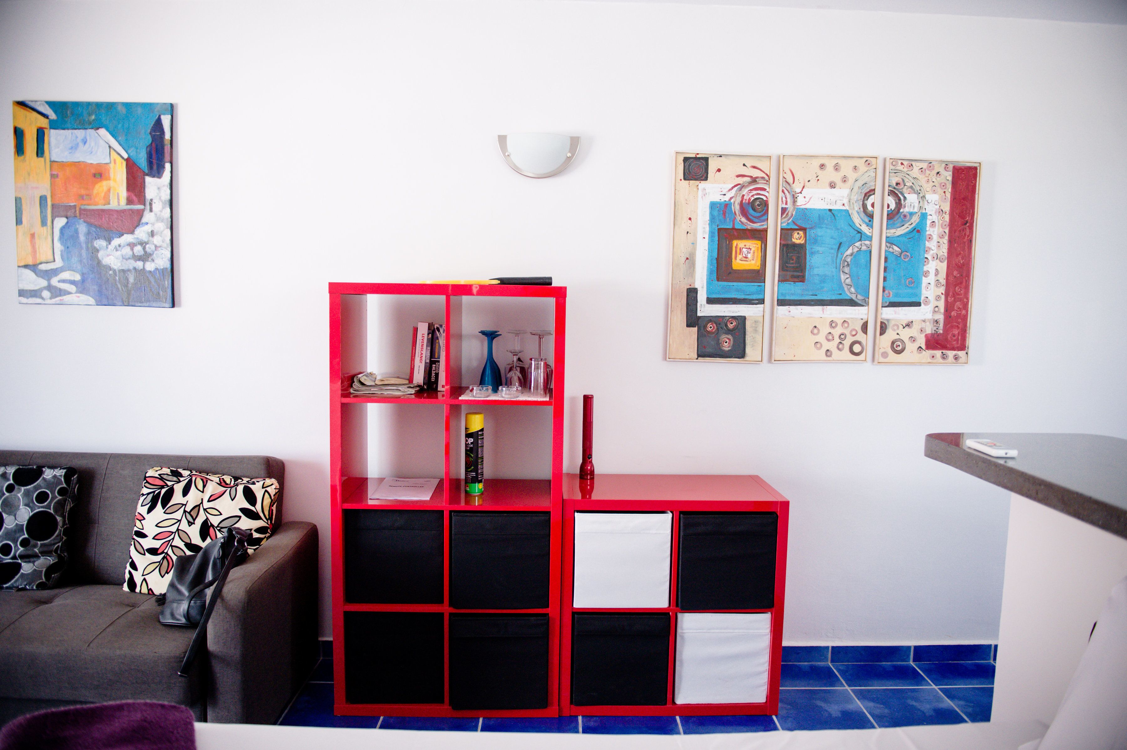 A living room with a red bookshelf, a grey sofa, and two abstract paintings on the wall.