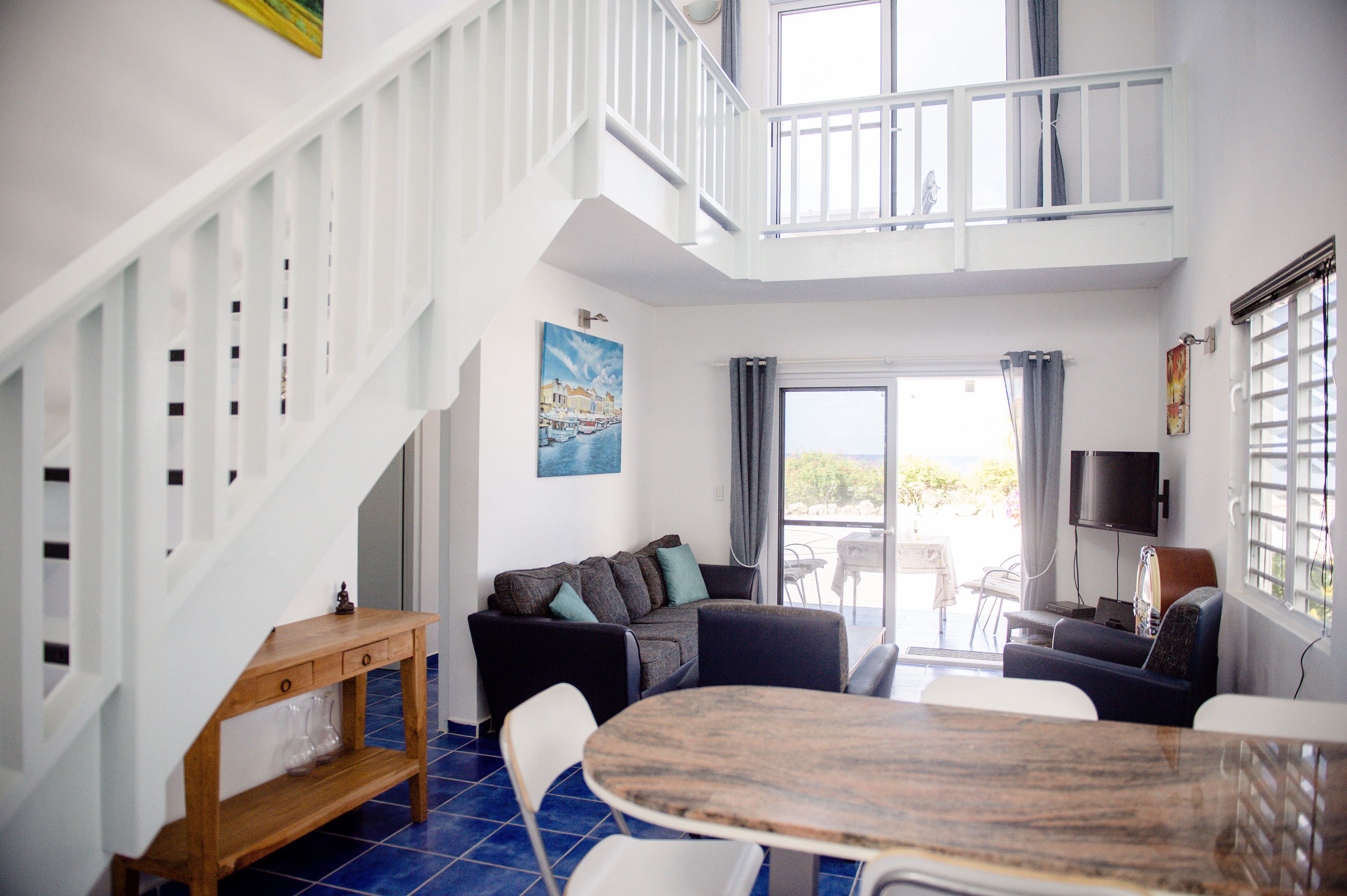 Uma sala de estar ampla com pé direito alto, escada de madeira, conjunto de sofá, mesa de jantar e televisão.