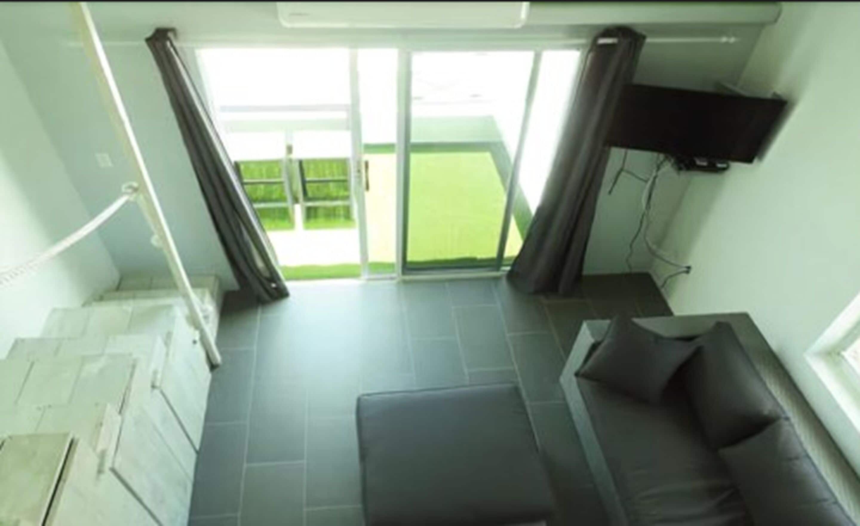 A room with a sofa, a TV mounted on the wall, and a view of a green outdoor area.