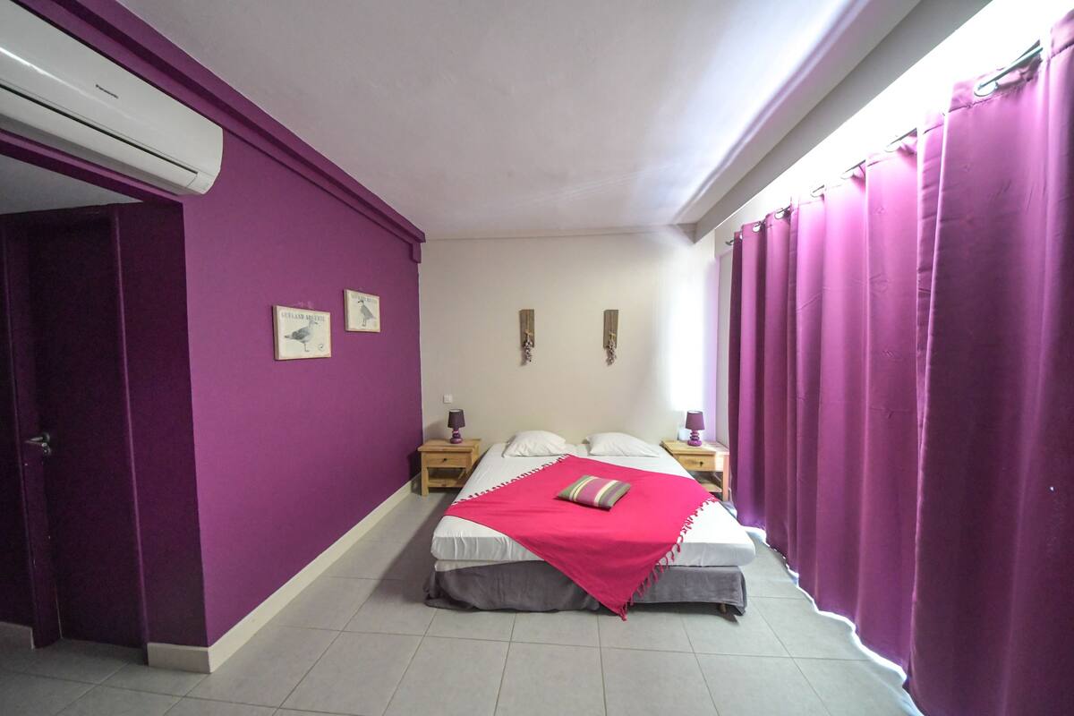 A bedroom with purple walls, a bed with white linens, two bedside tables with lamps, and a window with purple curtains.