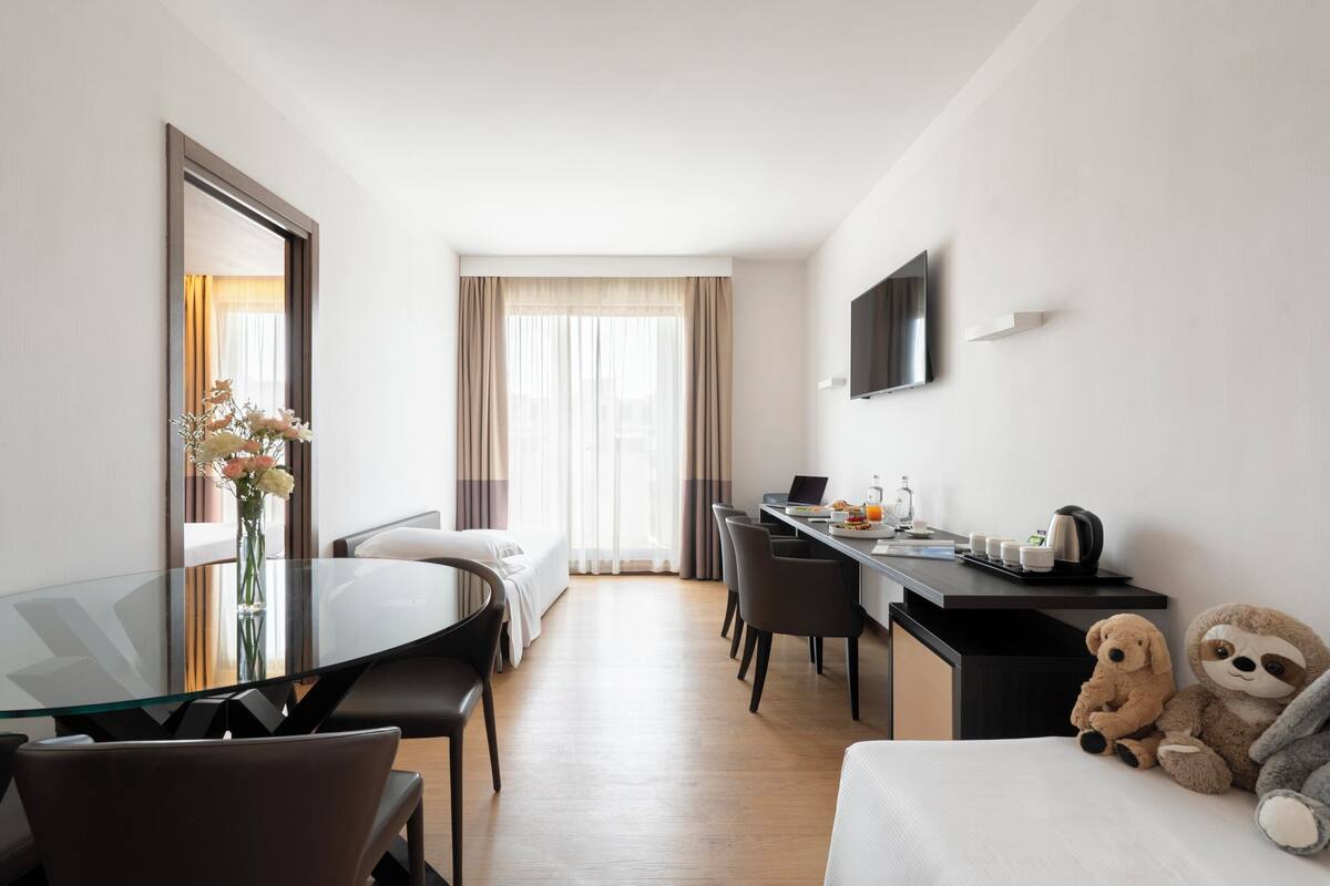 Una habitación de hotel con una mesa de vidrio, una cama, un escritorio con una computadora portátil, un televisor y peluches.