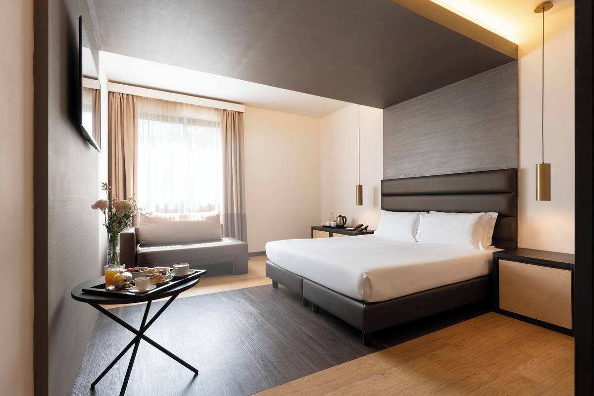 Una habitación de hotel moderna con una cama grande, mesitas de noche, una pequeña zona de estar y una bandeja de desayuno.