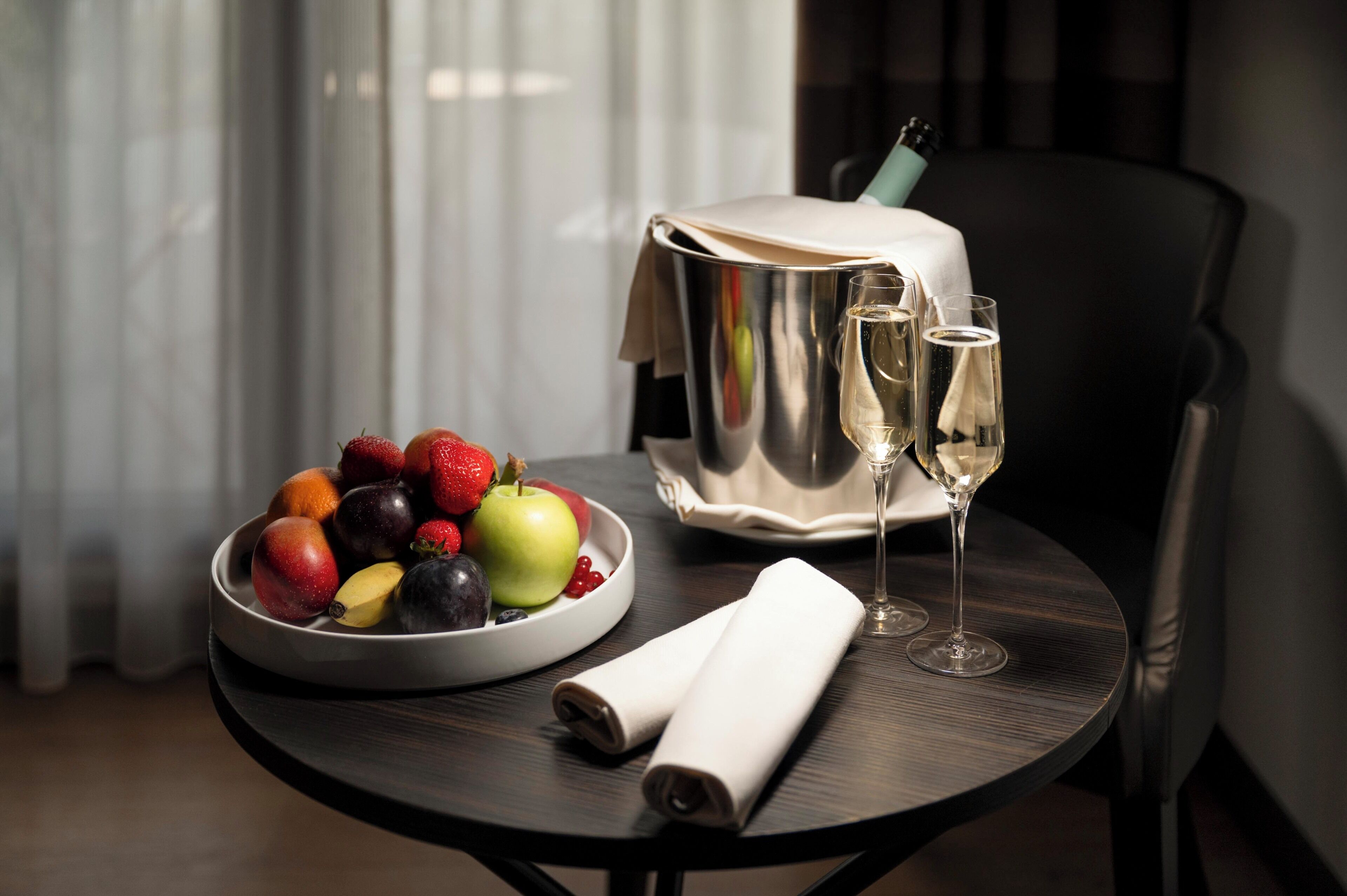Una mesa con un frutero, dos copas de vino, una botella de champán y una toalla enrollada.