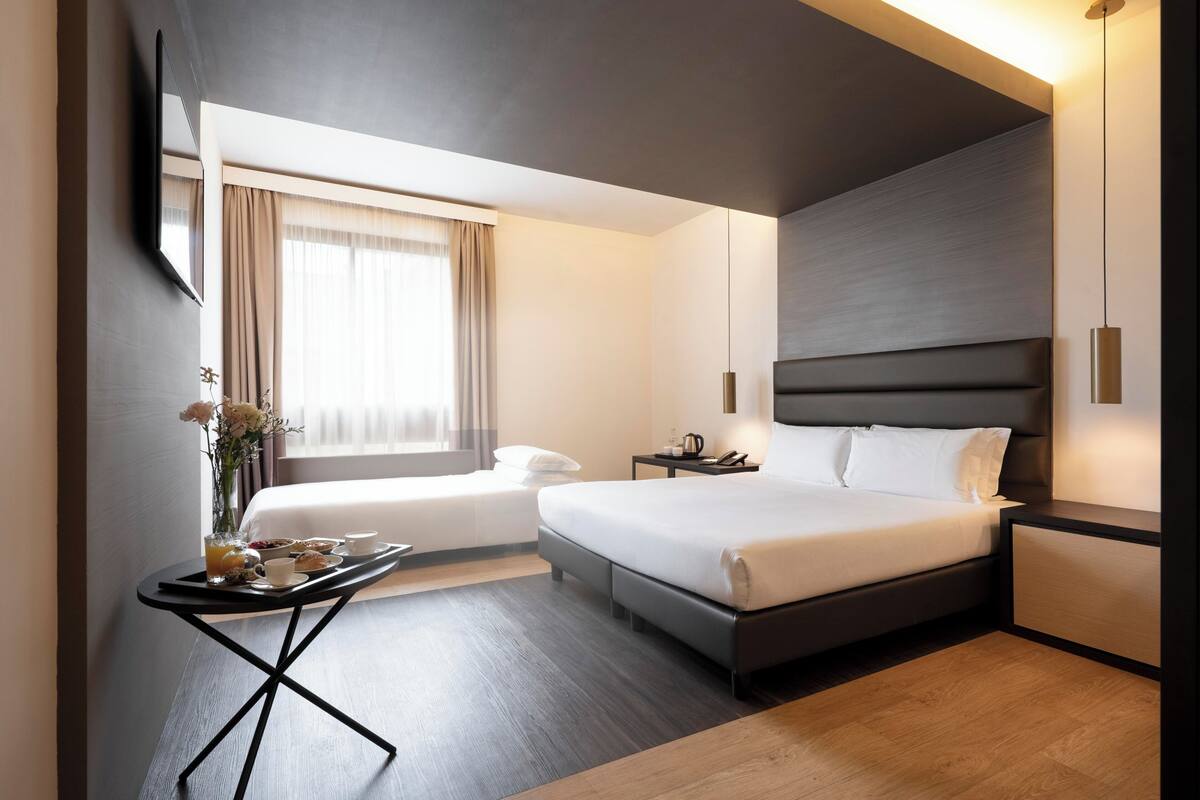 Una habitación de hotel moderna con una cama grande, mesitas de noche, una pequeña zona de estar y un ventanal con cortinas.