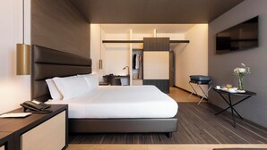 Une chambre d’hôtel moderne avec un grand lit, une table de chevet avec un téléphone, un petit bureau, une chaise, un téléviseur fixé au mur et une petite table sur laquelle est posé un vase de fleurs.