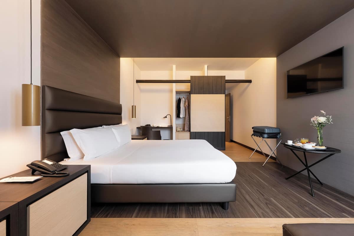 Una habitación de hotel moderna con una cama grande, una mesita de noche con teléfono, un escritorio pequeño, una silla, un televisor montado en la pared y una pequeña mesa con un jarrón de flores.