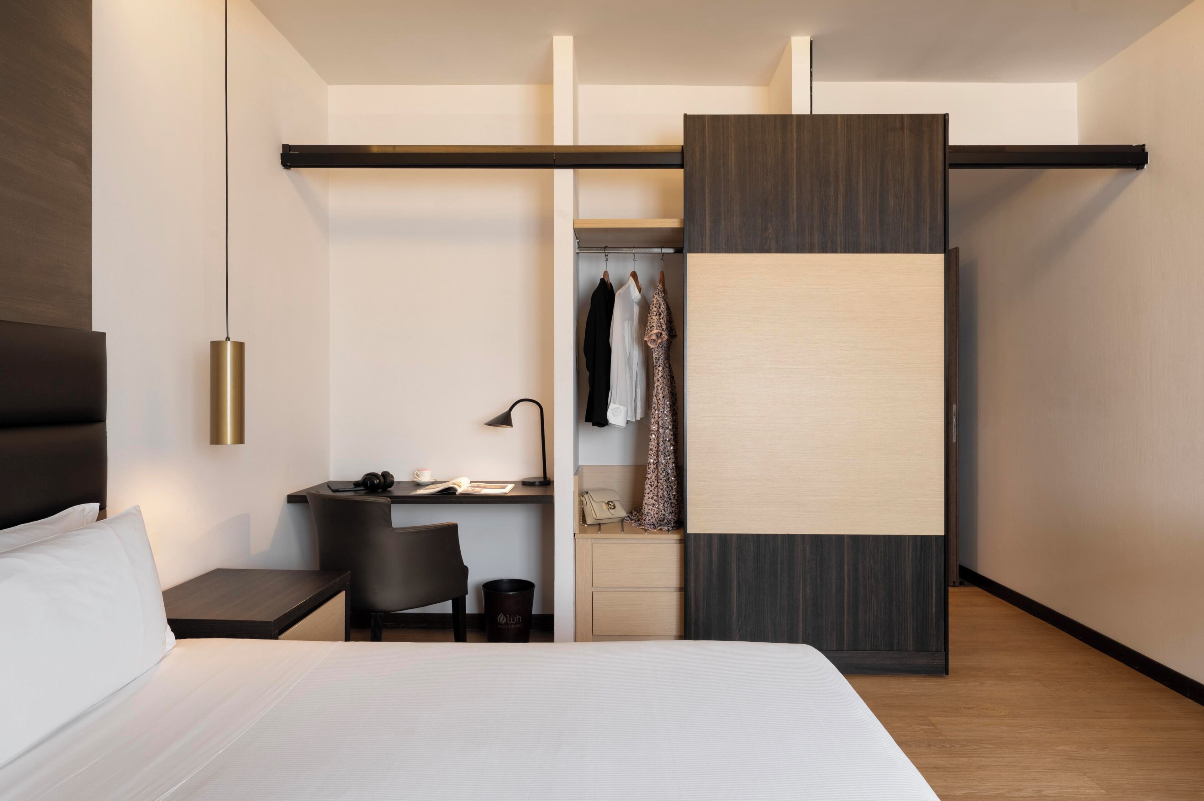 Quarto de hotel moderno com cama, escrivaninha, cadeira, guarda-roupa e cabideiro.
