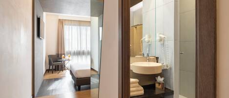 Una habitación de hotel moderna con cama, escritorio, baño con lavamanos y ducha, y un espejo.
