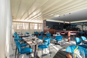 A restaurant with blue chairs, tables set for dining, a bar area, and large windows allowing natural light.