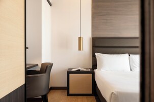 Habitación de hotel con cama, escritorio, silla, mesita de noche con un libro y una lámpara colgante.