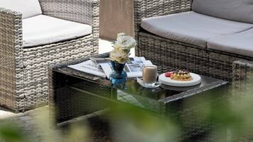 Un conjunto de sofá de mimbre con cojines, una mesa de centro con un florero y un plato de comida, y un camino de piedra que conduce a una fuente de agua.