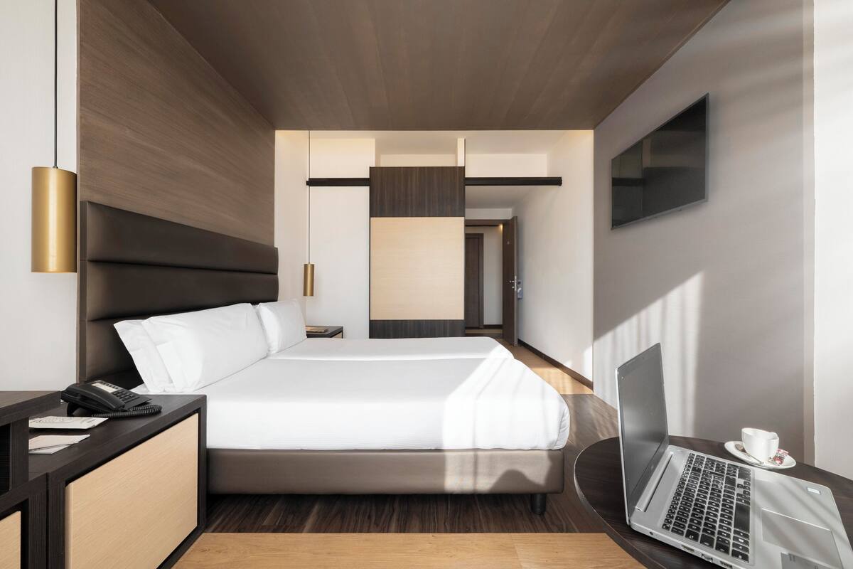 Una habitación de hotel moderna con cama, mesita de noche, lámpara, computadora portátil y televisor en la pared.