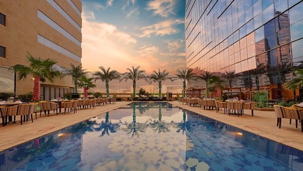 Uma área de piscina de hotel com piscina azulejada, palmeiras e mobiliário de exterior.