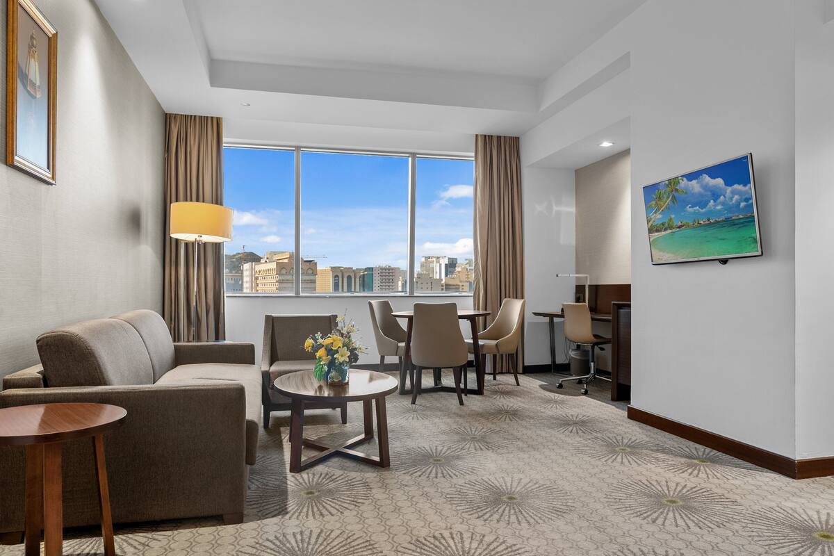 A hotel room with a large window, a sofa, a dining table with chairs, a small coffee table, and a TV on the wall.