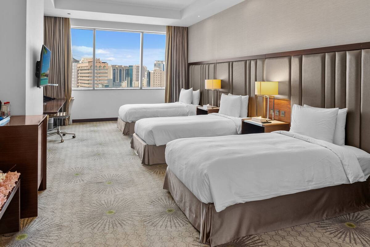 A hotel room with two beds, a desk, a TV, and a city view.