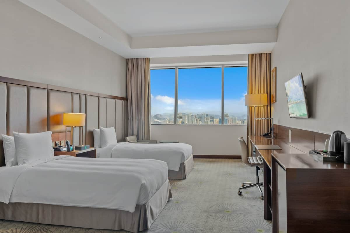 A hotel room with two beds, a desk, a chair, a TV, and a large window with a city view.