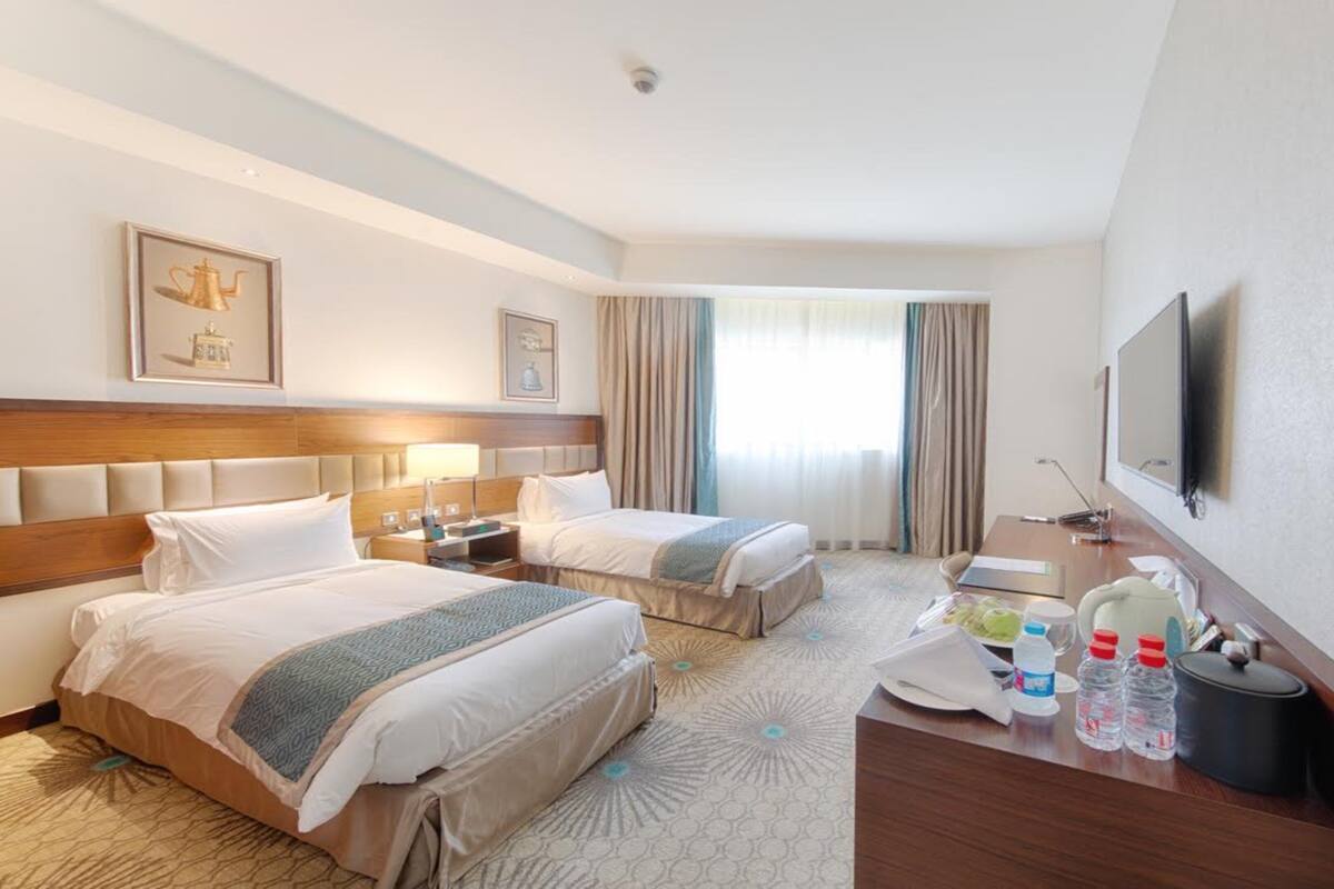 A hotel room with two beds, a desk, a TV, and a window with curtains.