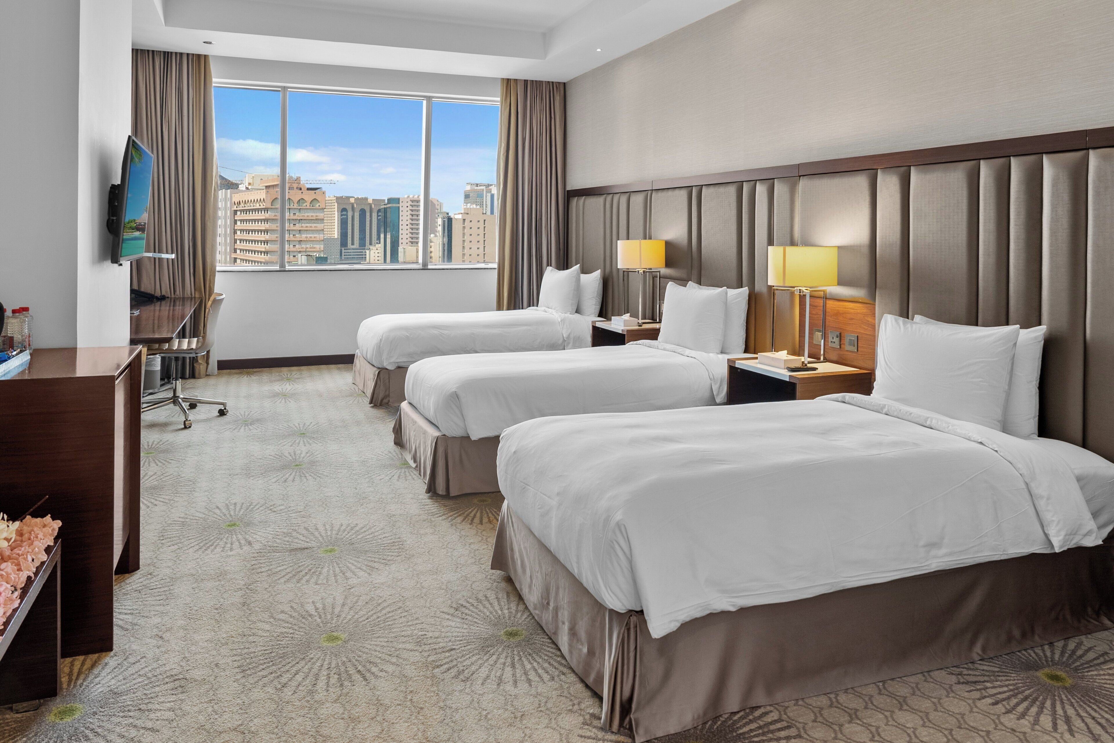 A hotel room with two beds, a desk, a TV, and a city view.