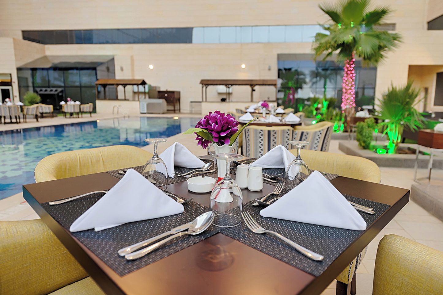 A dining table set for four with a pool view, including chairs, tableware, and a vase with flowers.