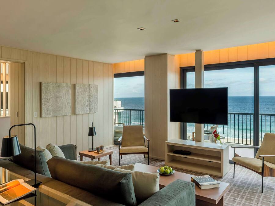 Una sala de estar moderna con un televisor grande, asientos cómodos, una mesa de centro y vistas al océano a través de amplias ventanas.