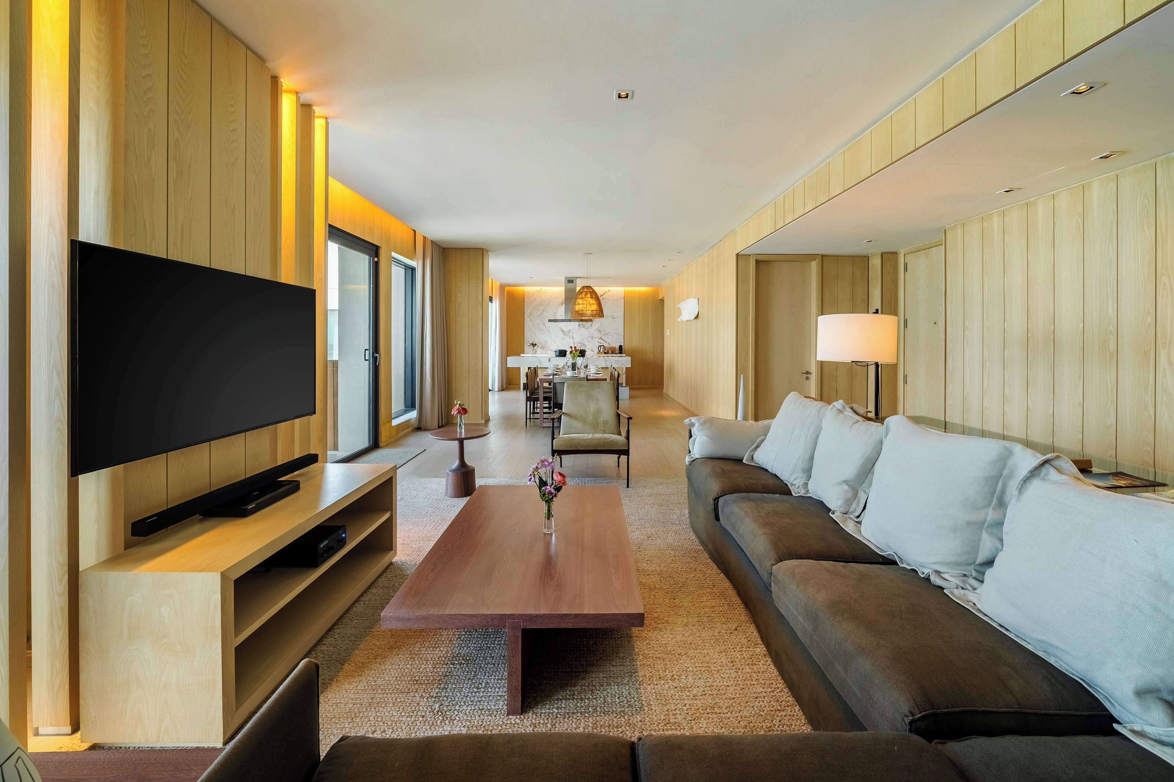 A modern living room with a flat-screen TV, a wooden coffee table, a sofa, and a dining area in the background.