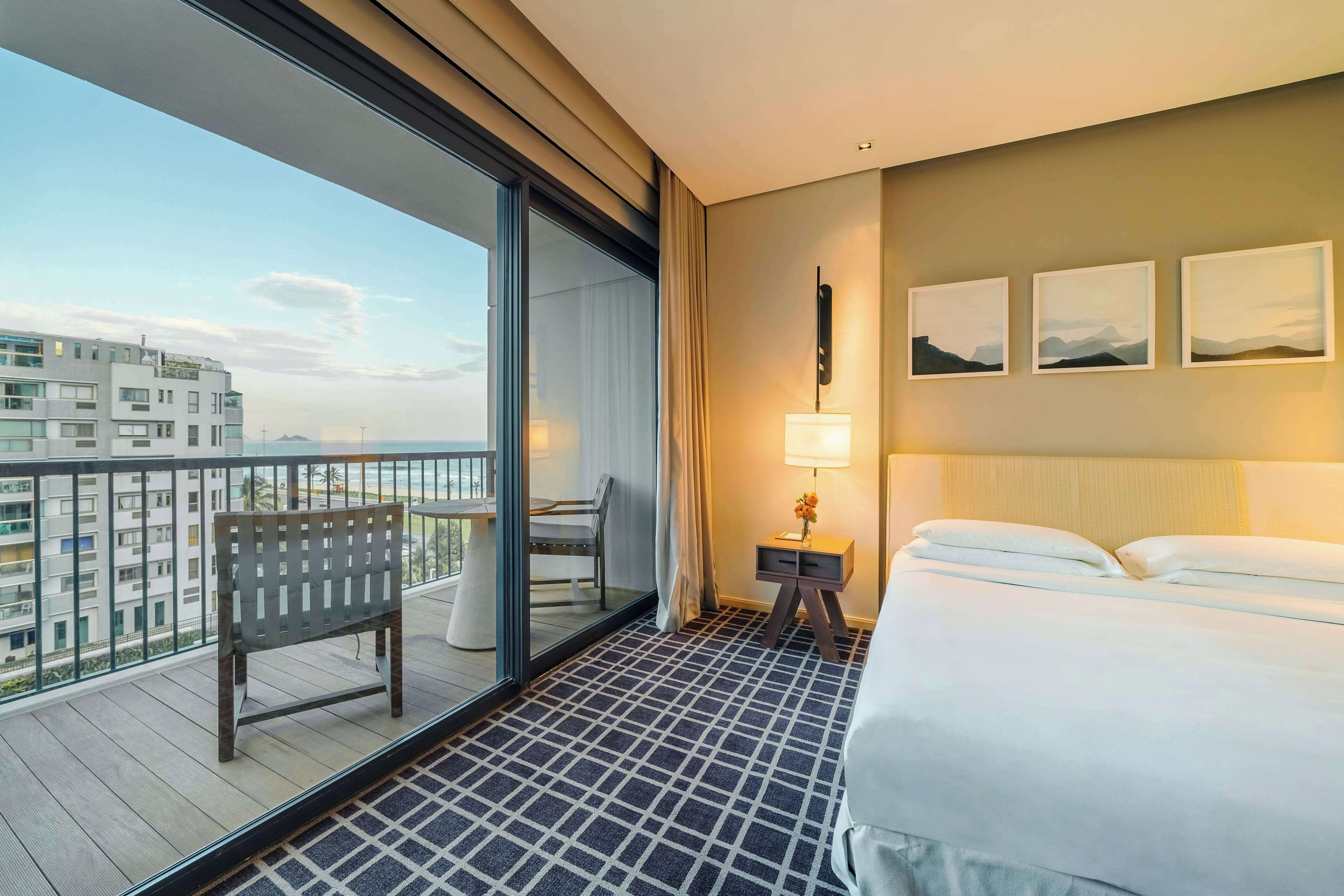 A hotel room with a bed, a chair, a small table with a lamp, and a balcony with a view of buildings.