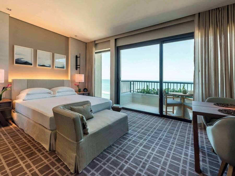 A hotel room with a large bed, a sofa, a desk, and a balcony with a view of the sea.