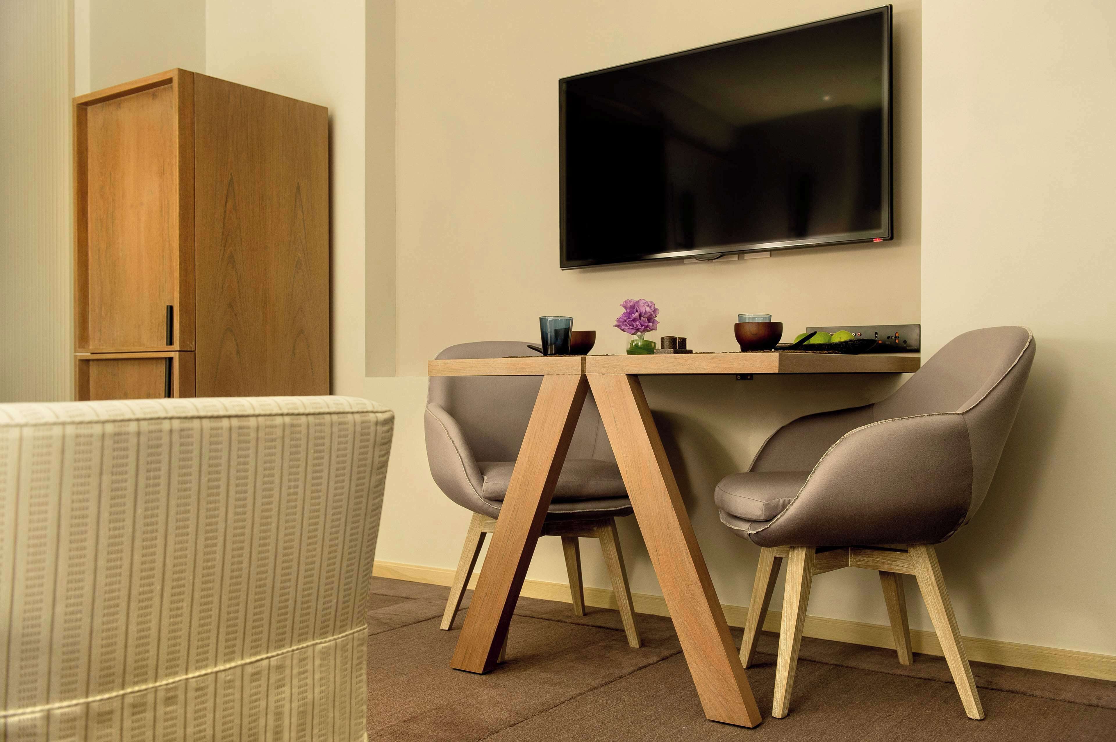A modern room with a wooden cabinet, a TV mounted on the wall, a dining table with two chairs, and a small vase with flowers.