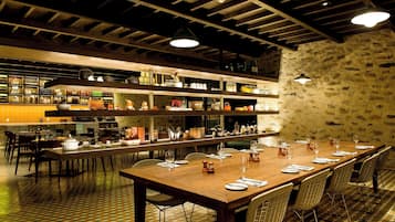 Un restaurante con una larga mesa de madera dispuesta para comer, varias áreas de comedor y una cocina con estantes llenos de diferentes artículos.