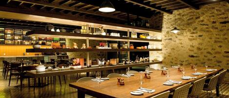A restaurant with a long wooden table set for dining, multiple dining areas, and a kitchen area with shelves stocked with various items.