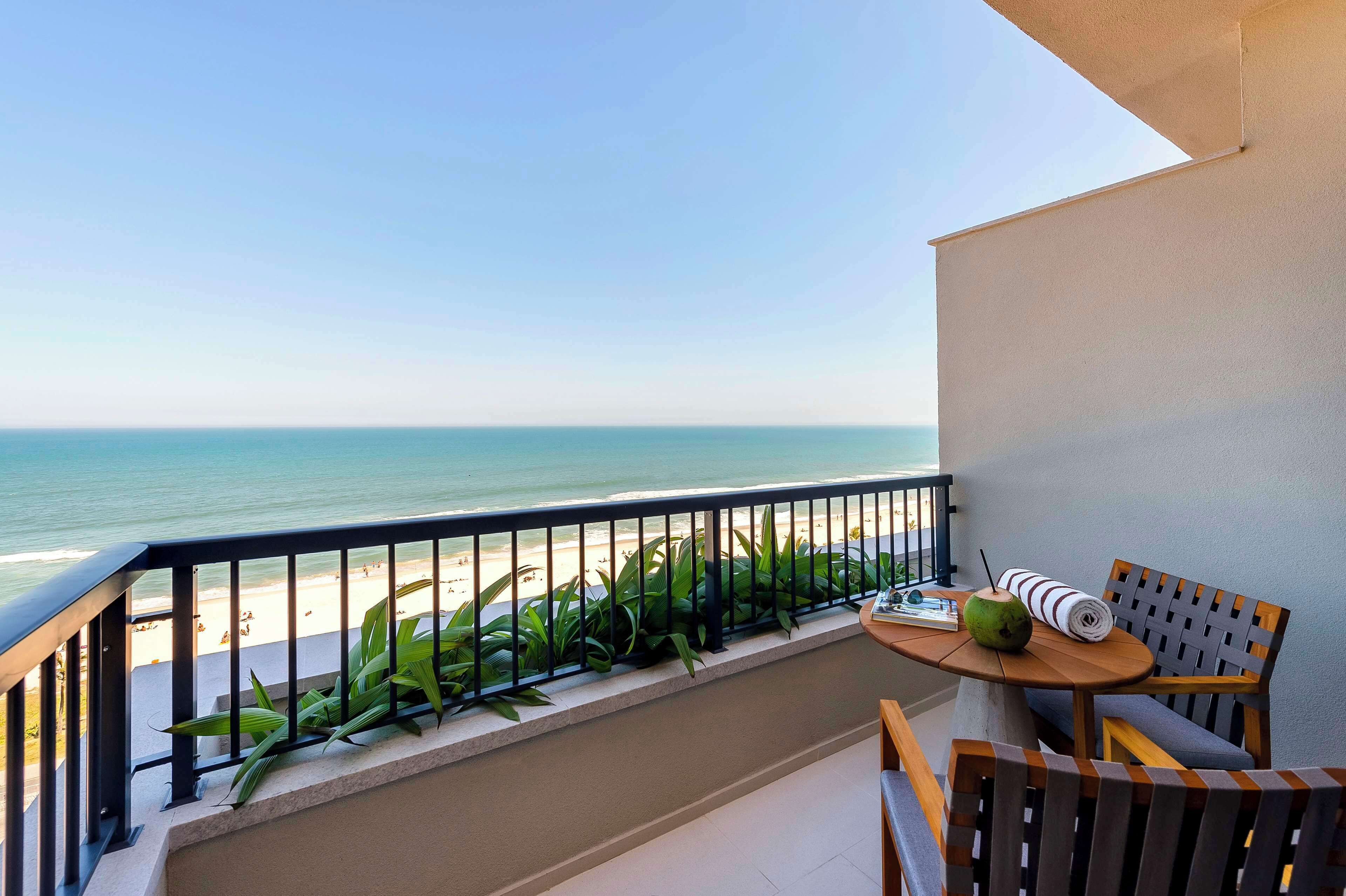 Un balcon donnant sur la plage, une table avec une noix de coco et une serviette enroulée, et une chaise.