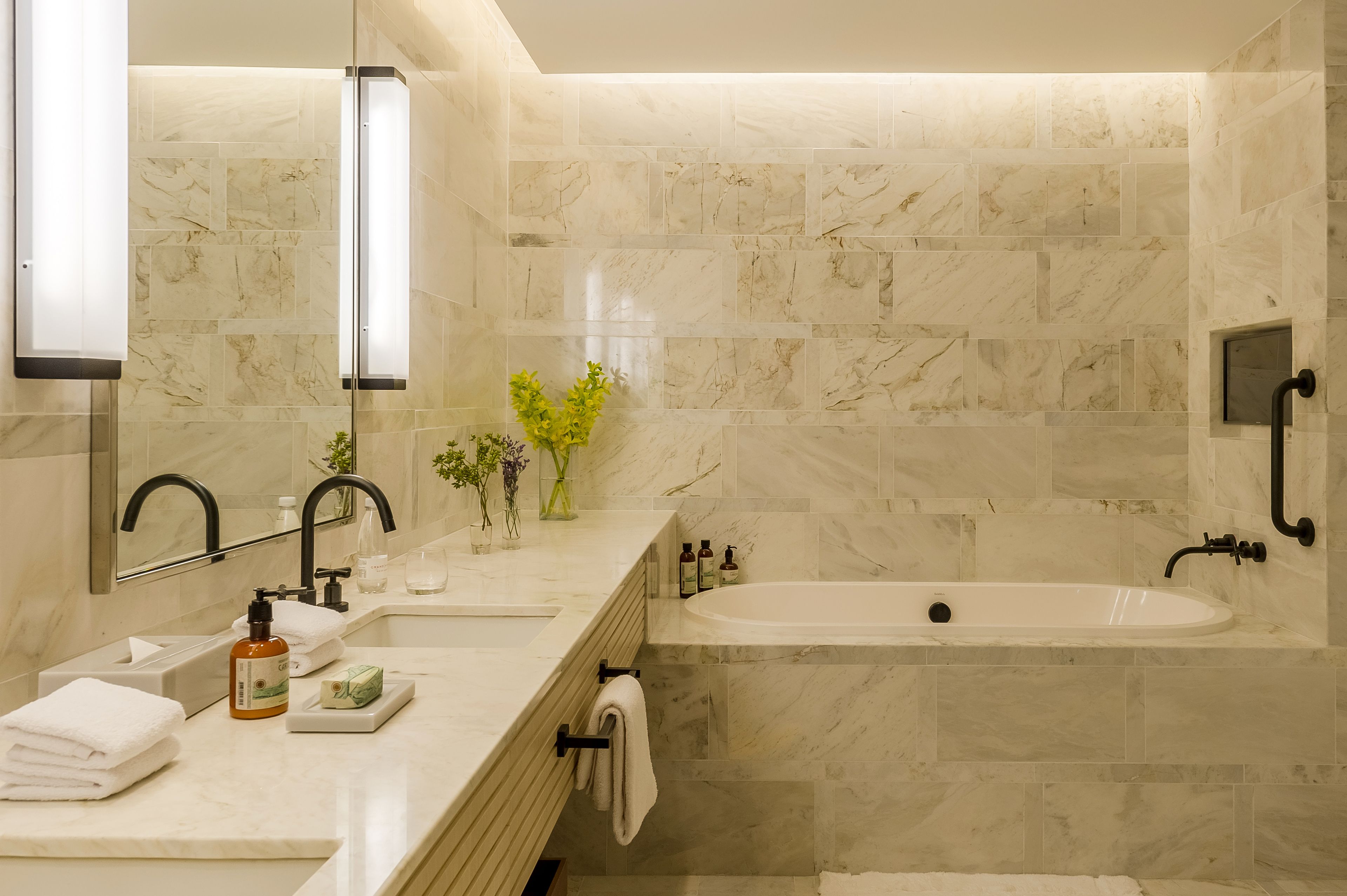 A modern bathroom with a marble tile wall, a large bathtub, a vanity with a mirror, and a sink with toiletries.