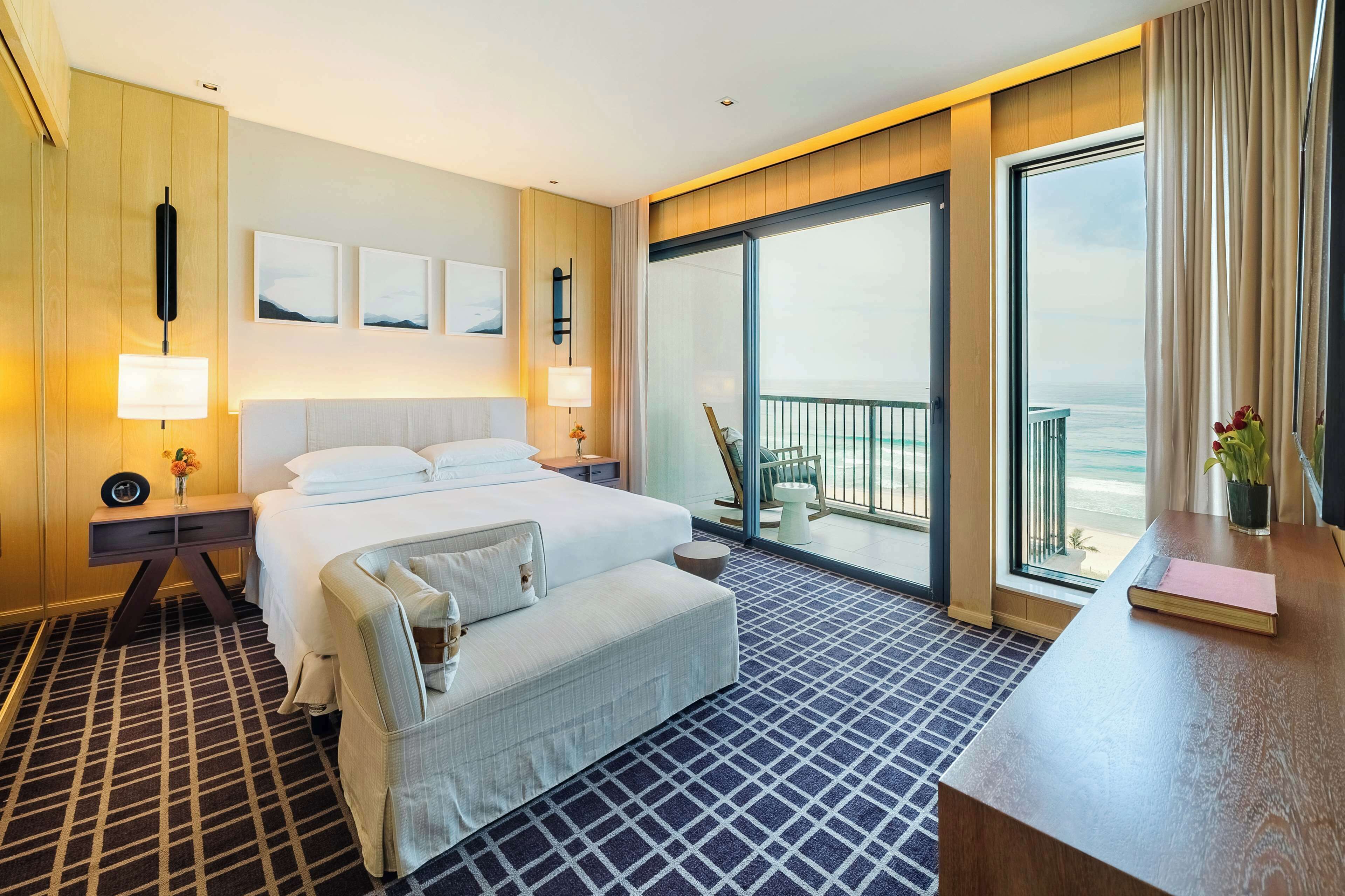 A hotel room with a large bed, a desk, a balcony with a view of the ocean, and a large window.