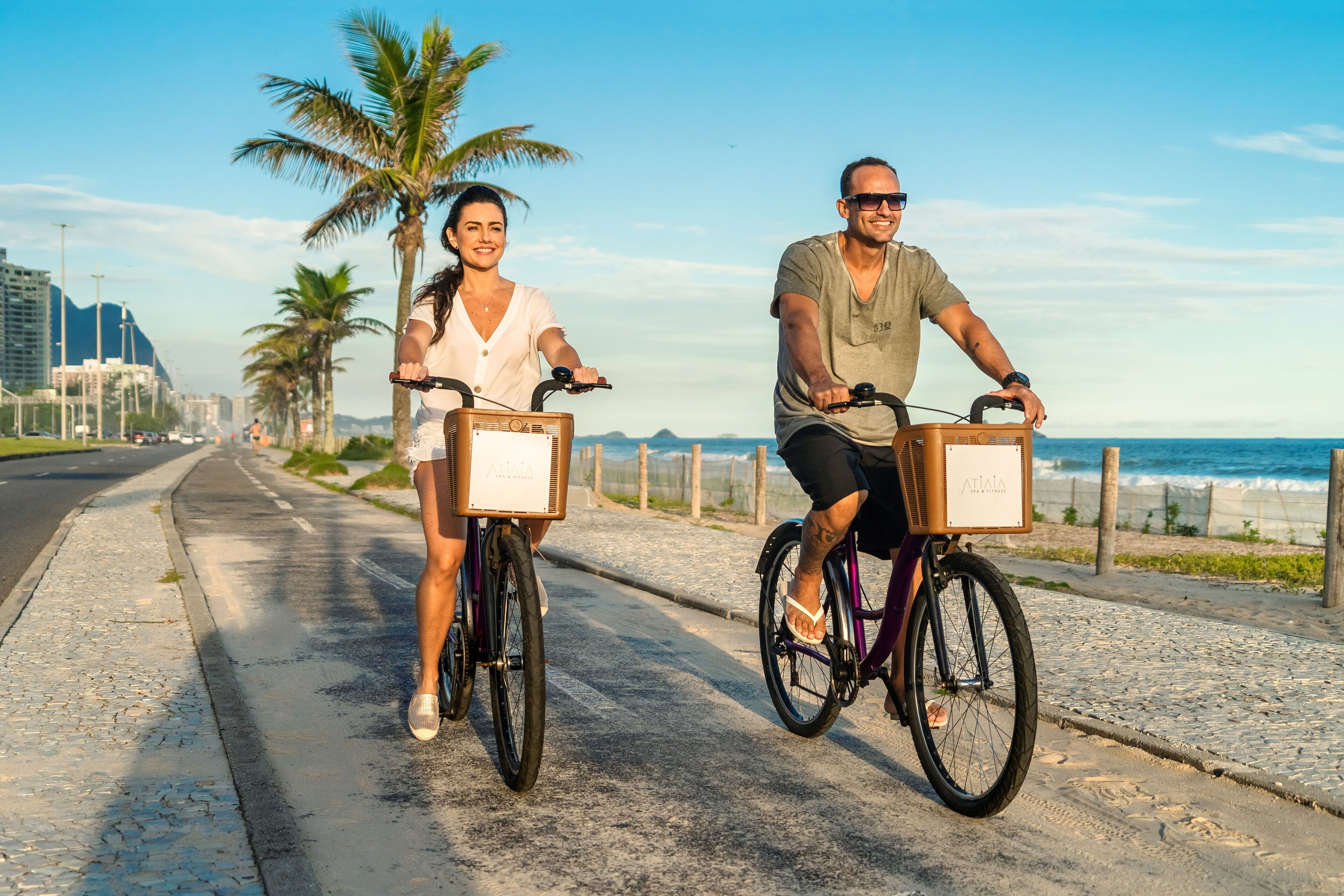 Two people riding bicycles on a paved path with a beach and palm trees in the background.