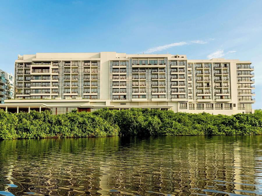 Um hotel moderno com vários andares e varandas, localizado à beira de um corpo d'água, com vegetação densa ao primeiro plano.
