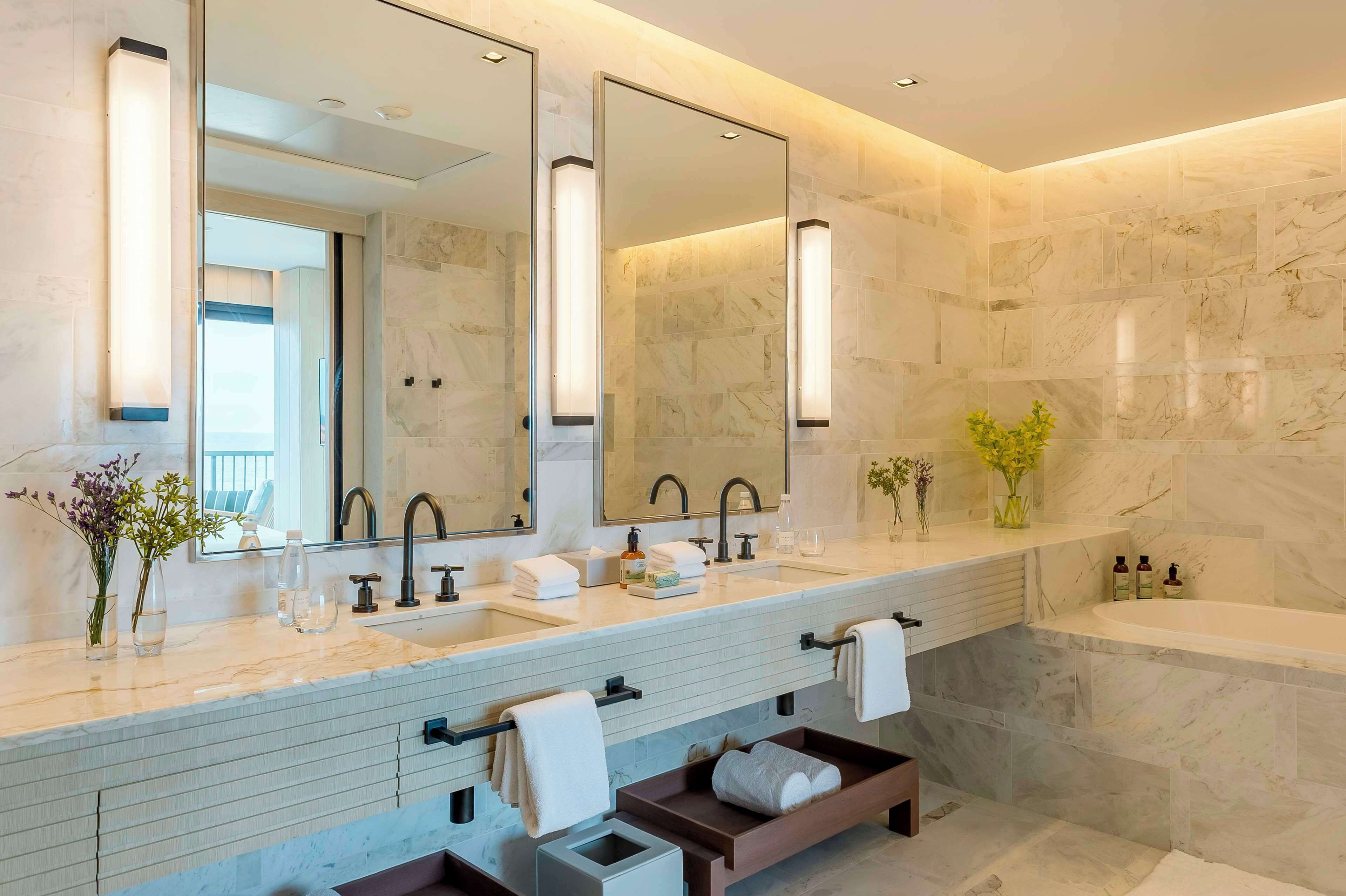 A modern bathroom with two sinks, a large mirror, and a bathtub. There are towels and toiletries on the counter.