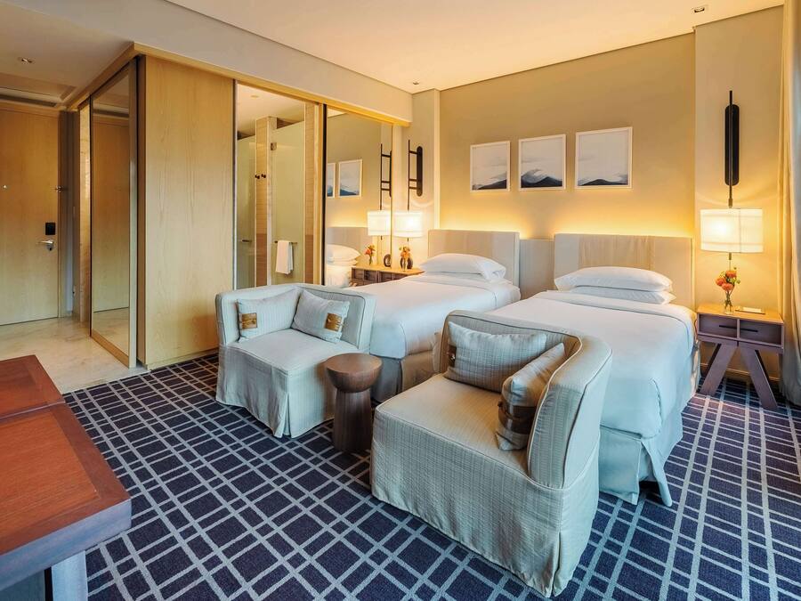 Quarto de hotel com duas camas, um sofá, uma poltrona, uma mesa de madeira e um tapete estampado.