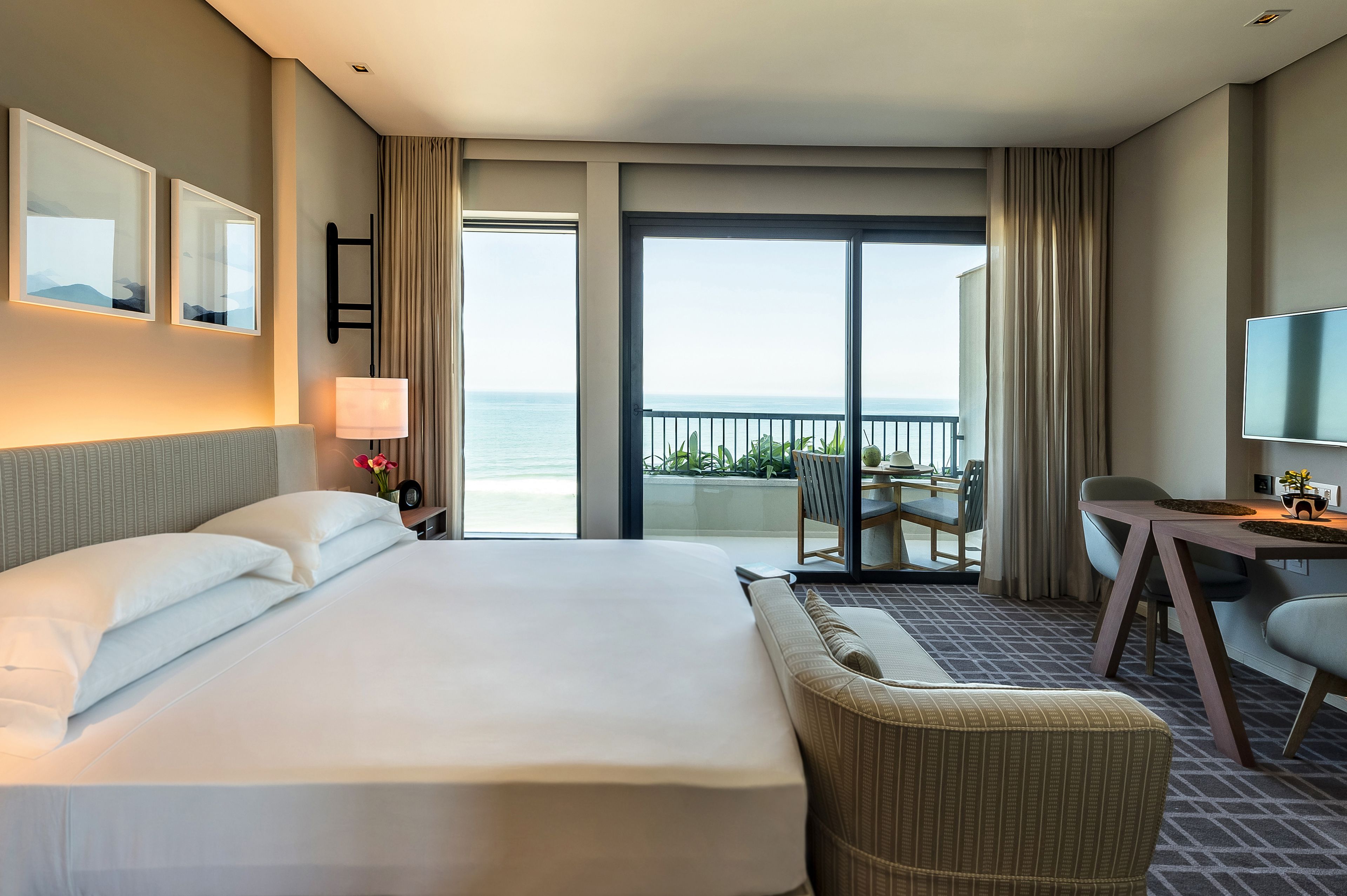 A hotel room with a large bed, a desk, a chair, a TV, and a balcony with a view of the ocean.